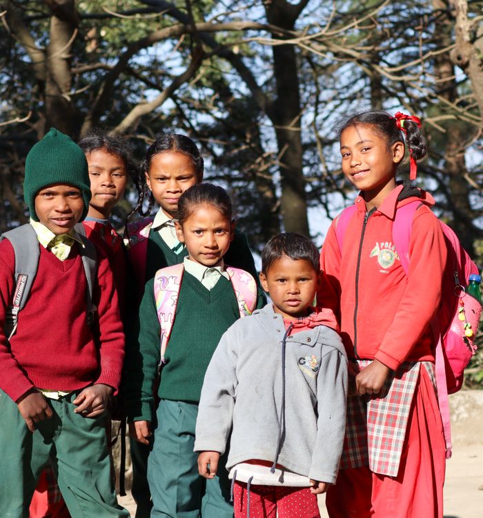 A group of smiling schoolchildren in uniforms and backpacks, standing outdoors in Kasauli, Himachal Pradesh.