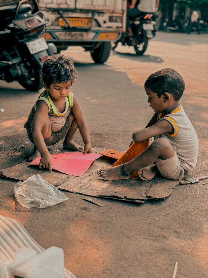 gallery-3 Two children playing creatively with paper on an Indian street, highlighting urban life and childhood innocence.