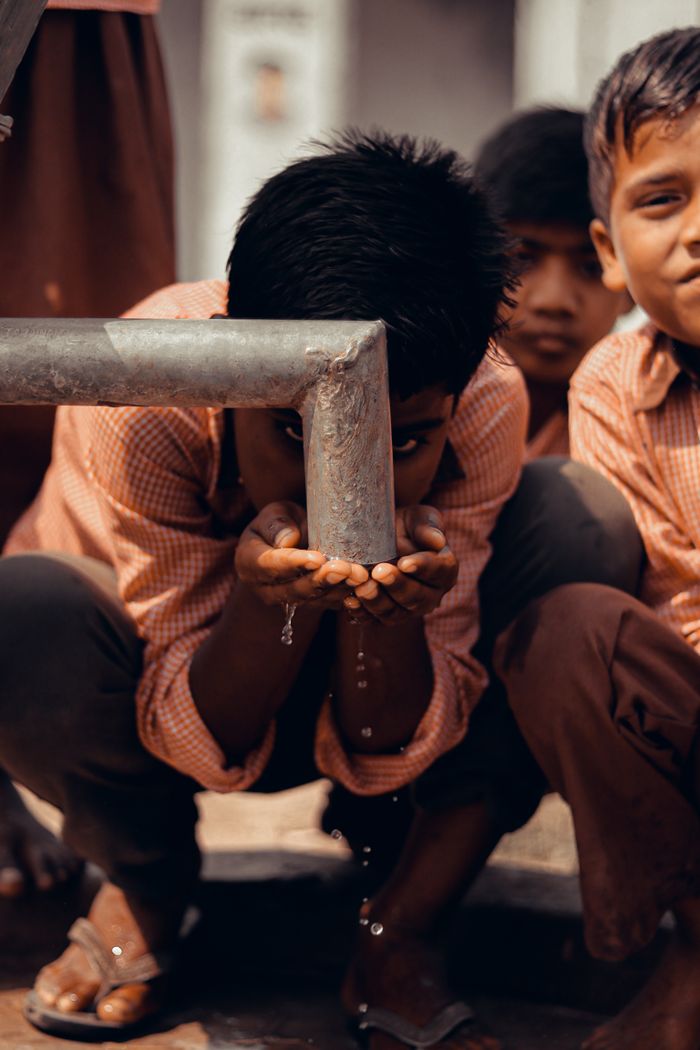 our-experience Ethnic boys in similar clothes sitting on haunches and drinking water from pipe on town street