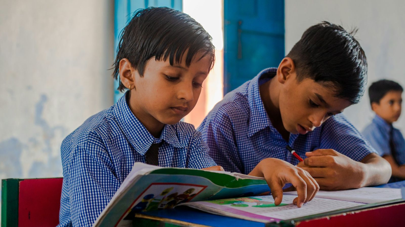 Two schoolboys in uniform reading and writing at their desk in an Indian classroom