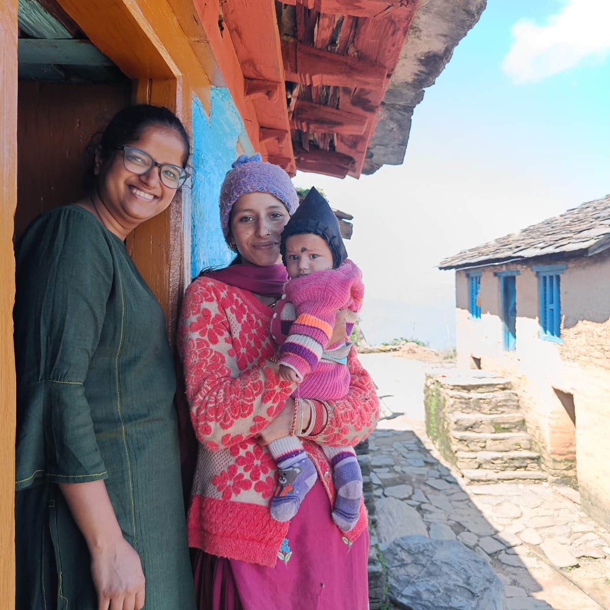 A smiling doctor with a mother and her baby outside a hillside home