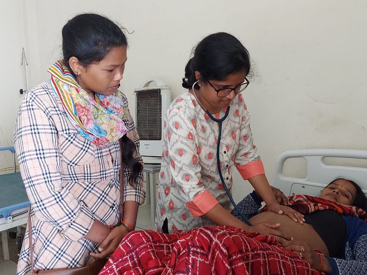 A female doctor examining a woman patient with a stethoscope while a young woman looks on