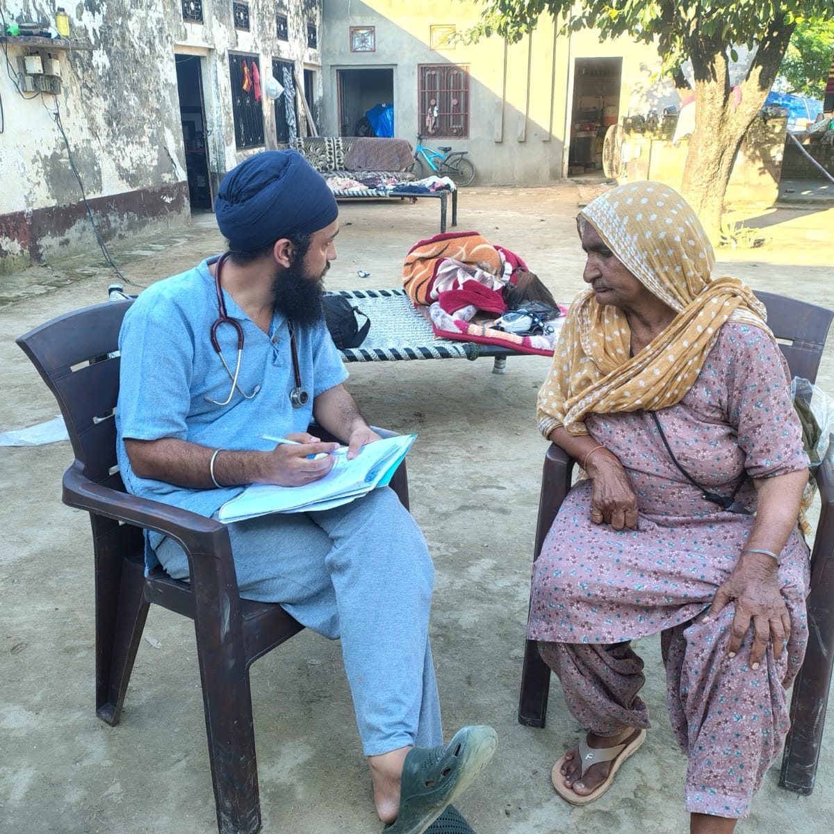 A doctor in scrubs taking notes while consulting an elderly woman in a village courtyard